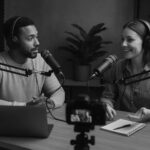 To personer sidder i et lydstudie med mikrofoner, hovedtelefoner og en bærbar computer foran sig, mens de optager en podcast og smiler under samtalen.